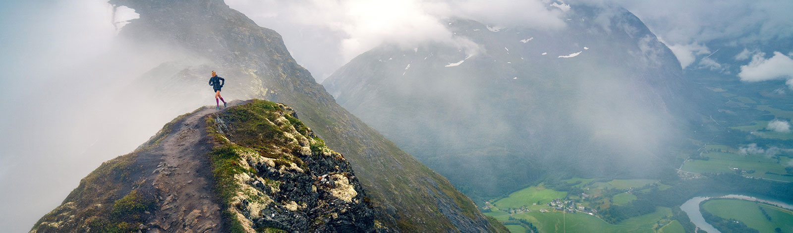 Neblige Gebirgslandschaft	 links läuft eine Frau über einen schmalen Bergpfad in der Nähe des Gipfels	 rechts ist das Tal mit einem kleinen Ort und einem Fluss zu erkennen 