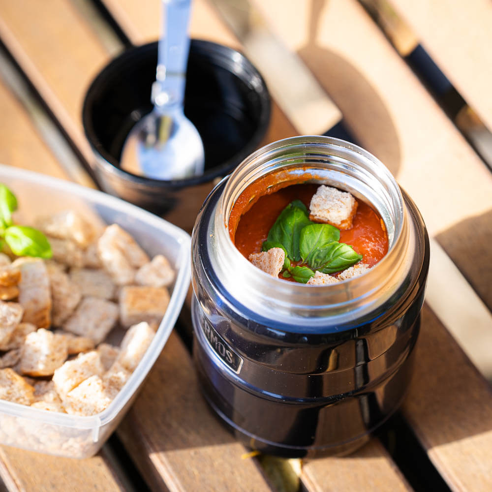 Wärmende Stärkung: Frische Tomatensuppe in der Thermoskanne, bereit für eine Pause Eine geöffnete Thermoskanne zeigt eine frische Tomatensuppe mit Basilikum und Croutons. Neben der Thermoskanne steht ein Behälter mit zusätzlichen Croutons und ein Löffel.