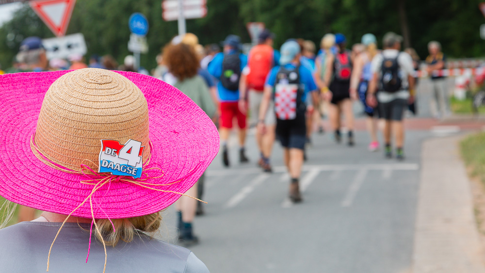 Eine Frau mit rosa farbenem Strohhut läuft eine Straße beim 4Daagse entlang. Vor ihr nur unscharf zu sehen sind andere Wanderer.