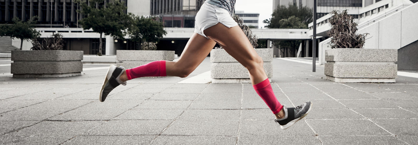 Durchbrechen das Grau: unsere Compression Sleeves in 7 Farben Frau mit pinken Wadensleeves läuft schnell und mit Beinen in der Luft durch eine urbane Betonwüste
