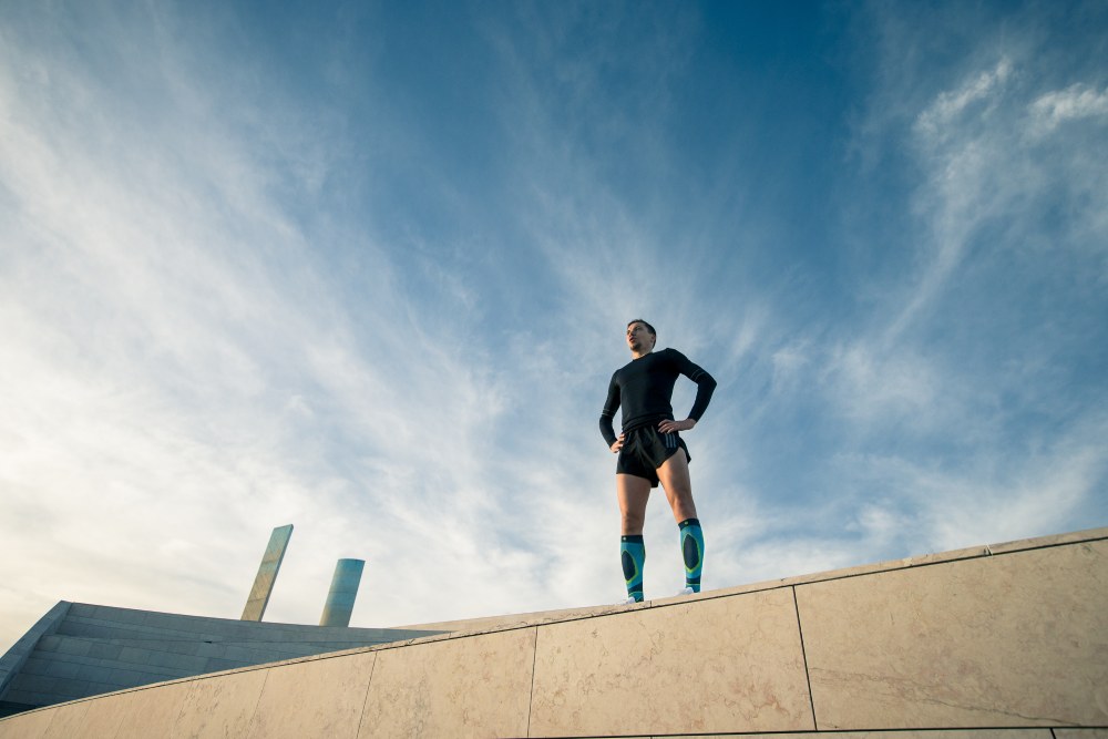 Läufer mit schwarzer Laufkleidung und blauen Laufsocken steht entschlossen auf der Mauer eines modernen Gebäudes und sieht in die Ferne