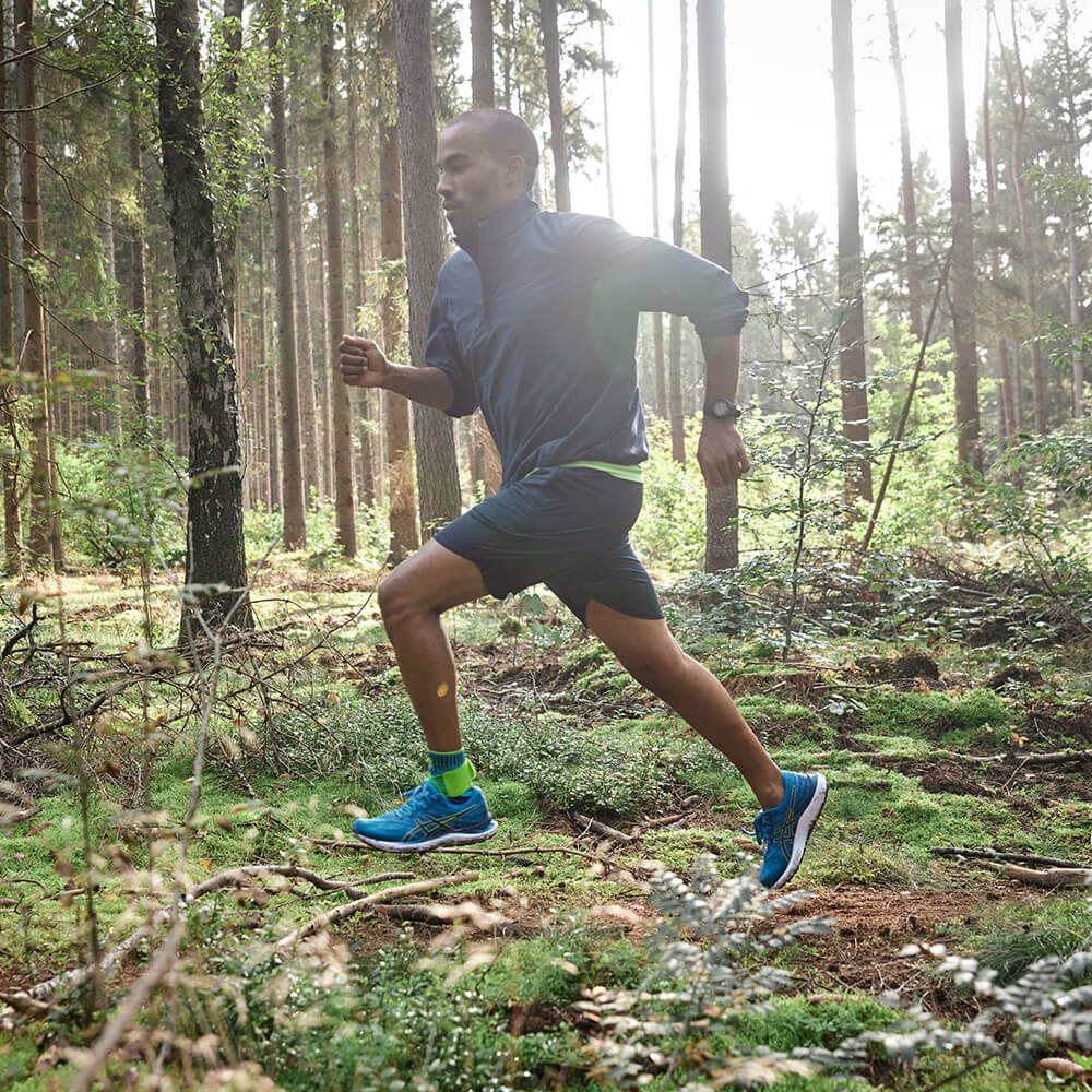 Lauf dich frei – mit stabiler Unterstützung durch unebenes Gelände. Ein Mann läuft dynamisch durch einen sonnigen Wald. Er trägt eine blaue Jacke, schwarze Shorts und leuchtend grüne Knöchelbandage.