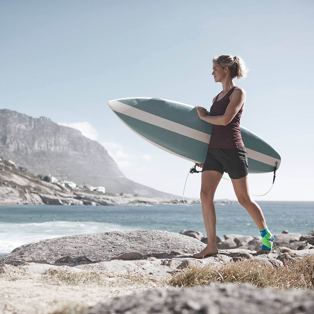 Frau mit einem Surfbrett unter dem Arm mit einer Sprunggelenkbandage am linken Bein an einem steinigen Strand mit Meer und Bergen im Hintergrund