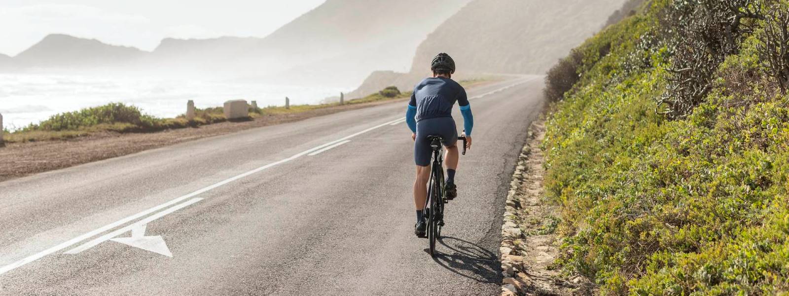 Fahrradfahrer mit Arm Sleeves auf Küstenstraße Radfahrer trägt blaue Arm Sleeves und fährt auf einer Straße am Hang mit Meerblick