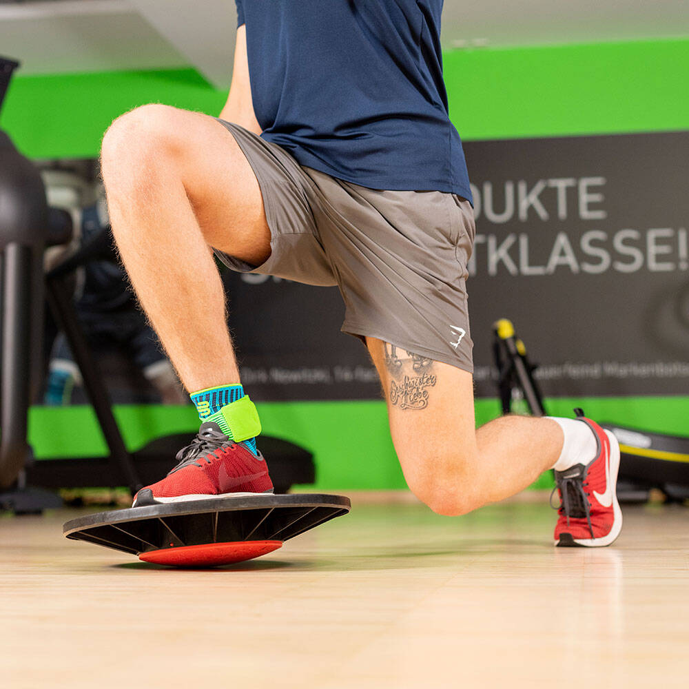 Lunges lassen sich mit einem Balance Pad prima zu einer komplexen Stabilisierungsübung erweitern Mann in einem Fitnessstudio macht einen Lunge mit dem vorderen Bein auf einem Balance Pad stehend und trägt eine Sprunggelenkbandage