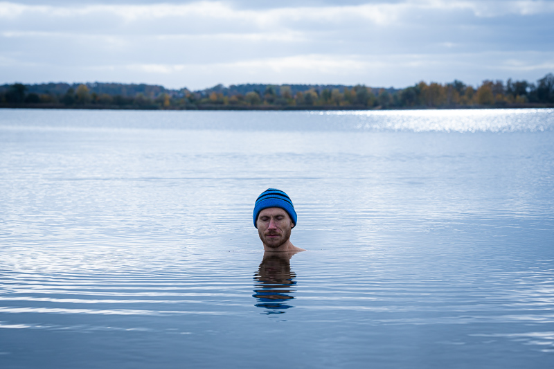 Eisbaden im See ist ein Hauch Abenteuer Ein Mann steht in einem ruhigen See, der bis zum Hals im Wasser ist. Er trägt eine blaue Mütze, und die Herbstlandschaft mit bunten Bäumen spiegelt sich im Wasser wider.