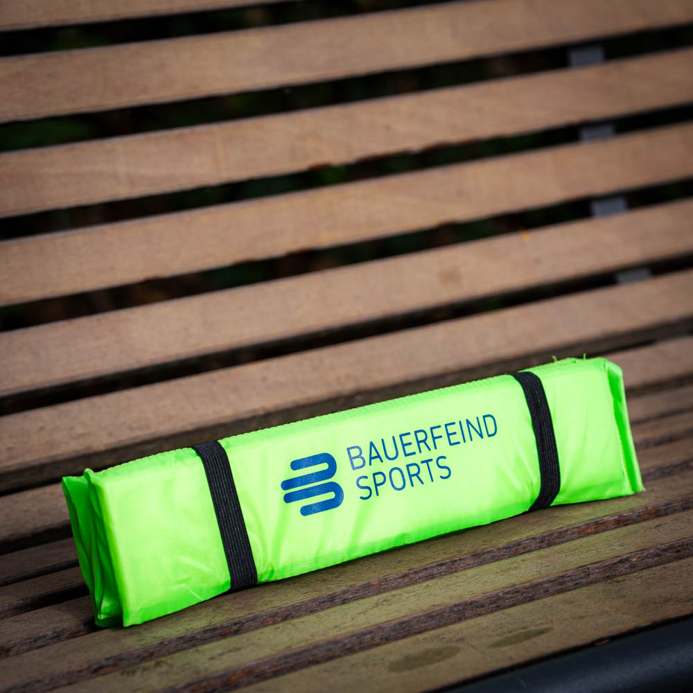 Praktisches Sitzkissen für unterwegs Gefaltetes Bauerfeind Sports Sitzkissen in leuchtendem Grün, sicher auf einer Holzbank verstaut.