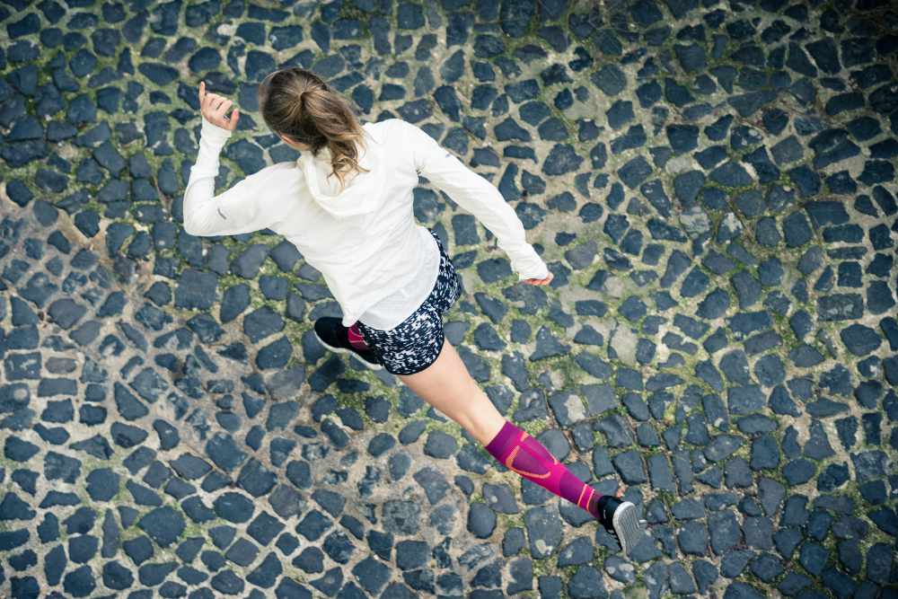 Vogelperspektive: Läuferin in weißer Laufjacke und mit rosa Laufsocken läuft über eine Altstadt-Straße