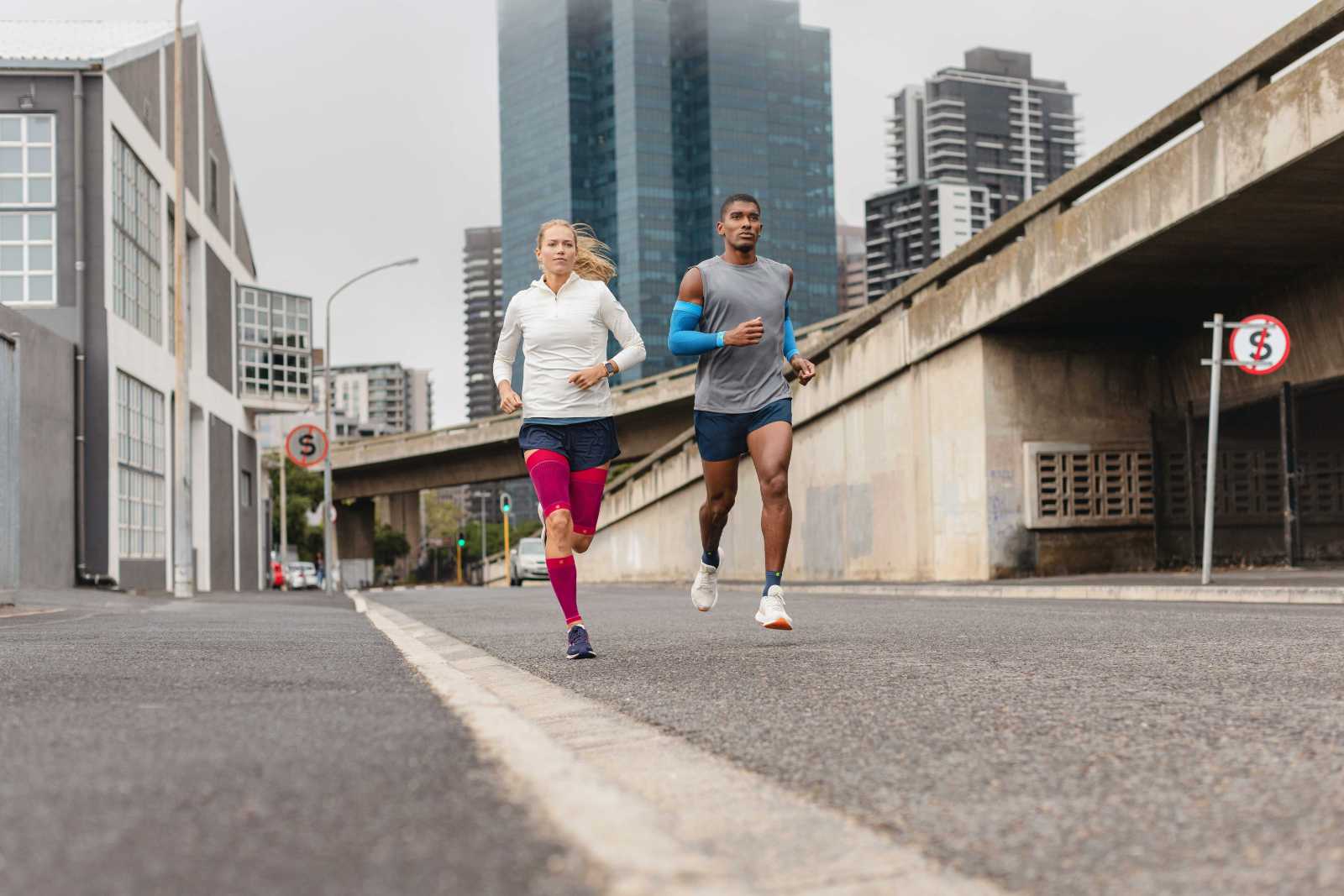 Eine Frau mit pinken Compression Sleeves und ein Mann mit blauen Arm Sleeves und kurzen Laufsocken laufen durch eine Großstadt