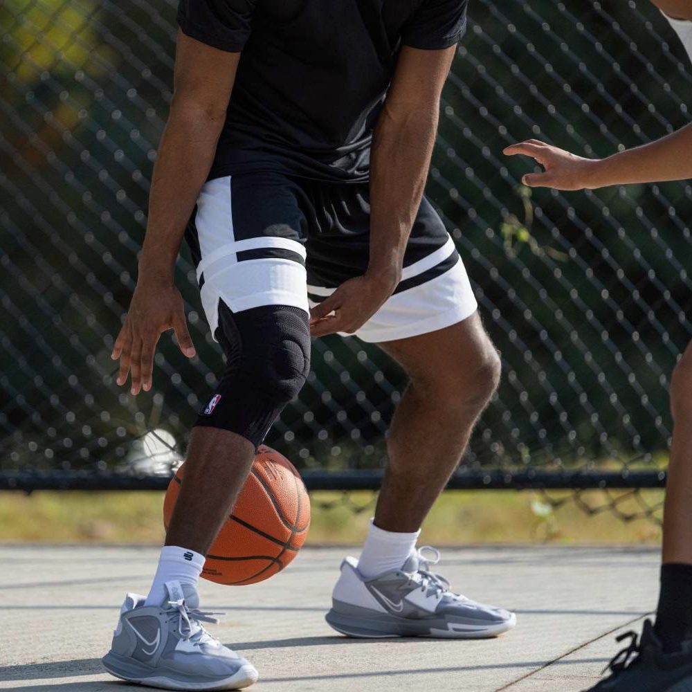 Gekonntes Dribbling mit schwarzer Kniebandage im Spiel Nahaufnahme eines Basketballspielers beim Dribbling auf dem Freiplatz. Am rechten Knie trägt er eine schwarze Kniebandage mit NBA-Logo. Ein Gegenspieler ist teilweise im Bild zu sehen.