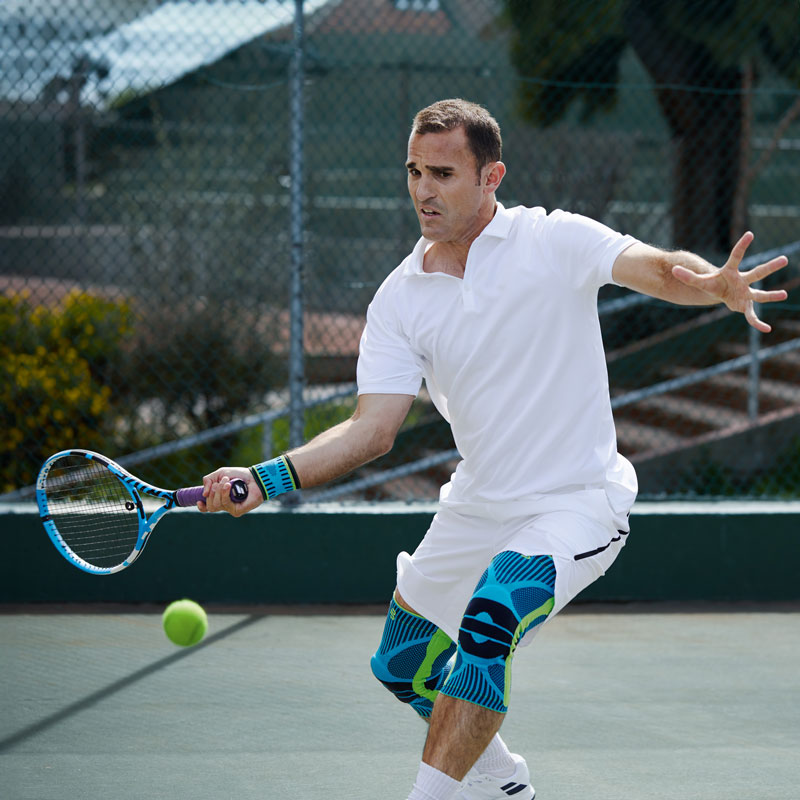 Tennisspielen mit Sports Knee Support und Sports Wrist Strap in rivera Tennisspieler mit weißer Tenniskleidung und Kniebandagen beim Vorhand Schlag auf einem Hartplatz