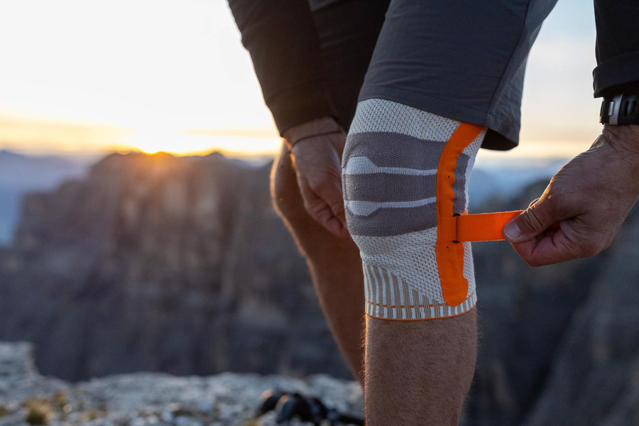 Ein Wanderer passt seine orangefarbene Kniebandage während einer Bergtour an. Im Hintergrund sind felsige Berge und ein Sonnenuntergang zu sehen.