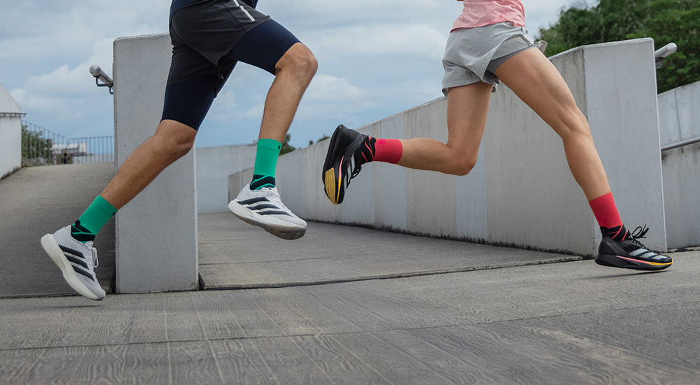 Mit Power nach vorne Zwei Läufer in Bewegung, die über eine Rampe sprinten. Der Mann trägt grüne Compression Socks und weiße Laufschuhe, die Frau schwarze Schuhe und rote Compression Socks.