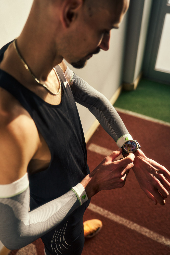 Hendrik Pfeiffer überprüft seine Laufuhr während des Trainings auf der Indoor-Laufbahn, mit Kompressions-Arm-Sleeves zur Unterstützung von Stabilität und Leistung.