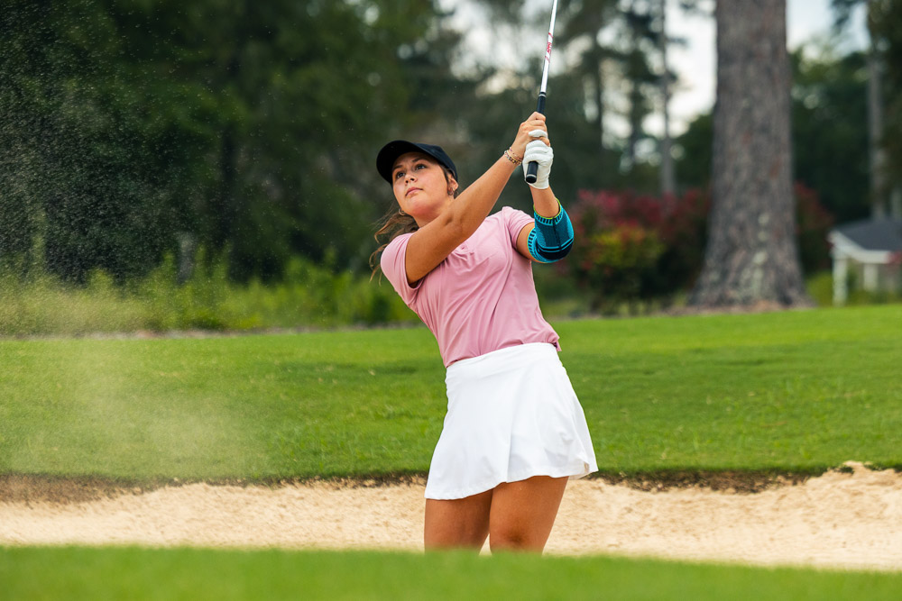 Golferin spielt aus dem Bunker und trägt eine Ellenbogenbandage für Unterstützung beim Golfschwung.
