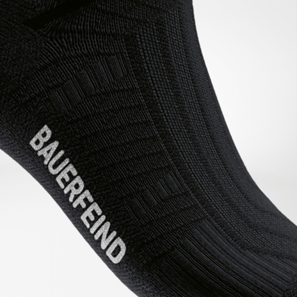 Seitlicher Blick auf eine schwarze Sportsocke mit gut sichtbarer, integrierter Rippstruktur am Mittelfußbereich und weißem „BAUERFEIND“-Schriftzug – unterstützt das Fußgewölbe beim Laufen.