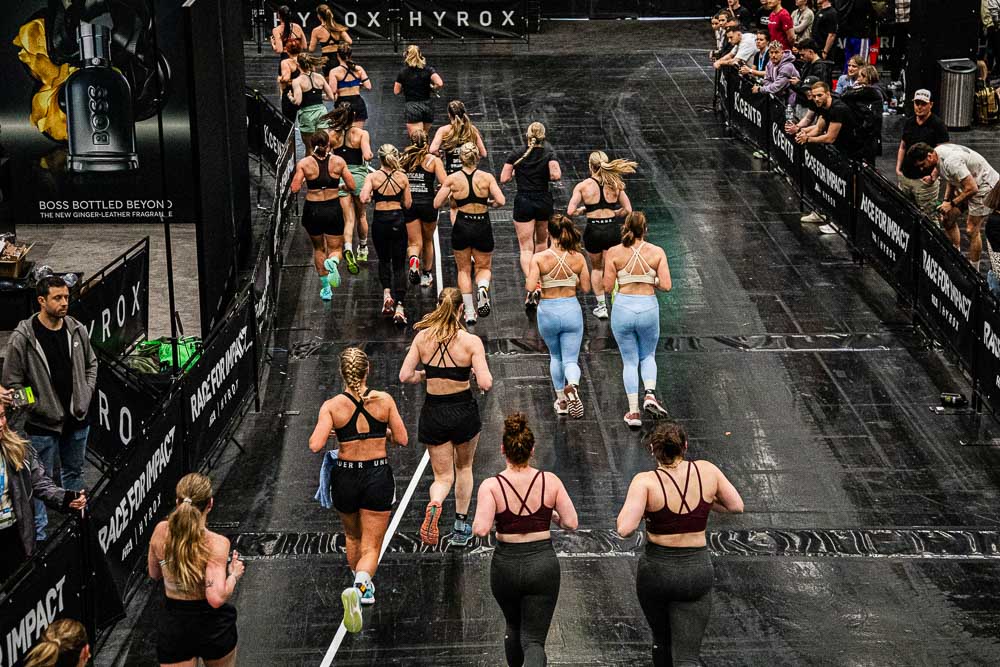 Blick von oben auf eine HYROX-Laufstrecke in einer Indoor-Eventhalle, auf der zahlreiche Athletinnen und Athleten in Sportkleidung unterwegs sind; Zuschauerinnen und Zuschauer stehen hinter den Absperrungen,