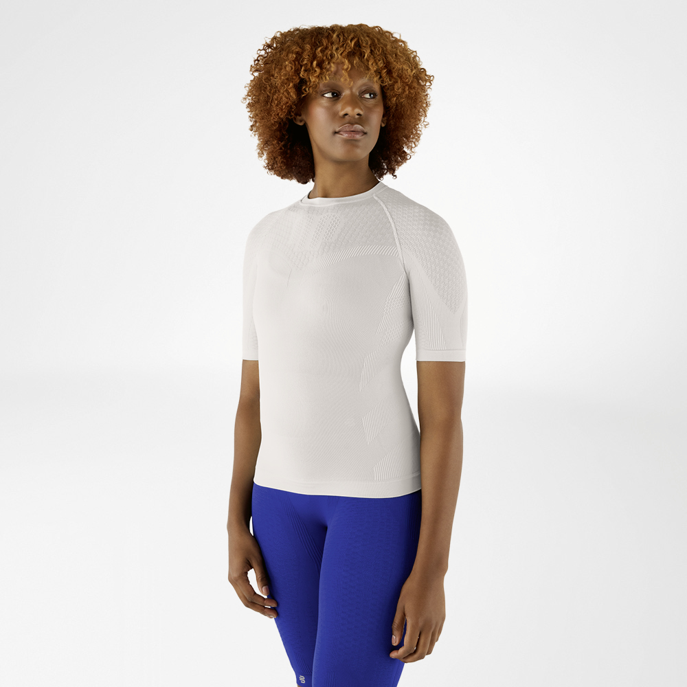 Frau mit lockigem Haar in einem silbergrauen Kompressionsshirt und blauen Leggings. Sie steht frontal und schaut zur Seite.