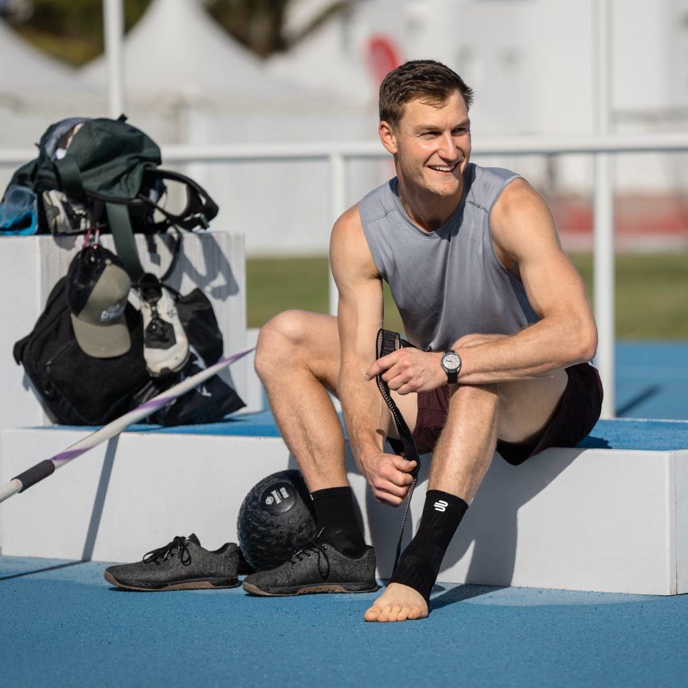 Bereit für den nächsten Sprung – mit stabilen Knöcheln! Ein Athlet sitzt auf einem Podest einer Leichtathletikanlage und zieht sich eine Bauerfeind Sports Ankle Support Bandage über den Knöchel. Neben ihm liegen seine Sportschuhe und eine Trainingskugel.
