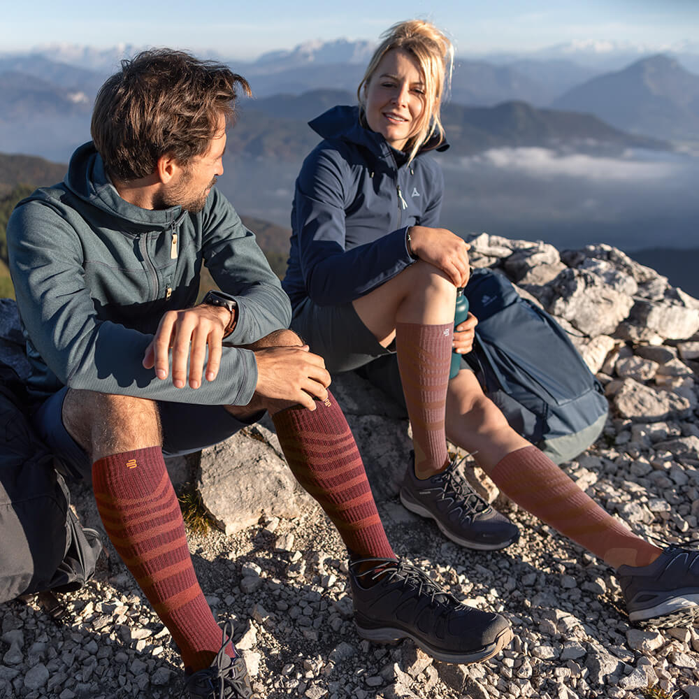 Zwei Wanderer genießen die Aussicht auf einem Berggipfel, während sie Merino Outdoor Kompressionssocken in Rot-Braun tragen. Die Socken bieten optimalen Komfort und Unterstützung für lange Touren in anspruchsvollem Gelände.