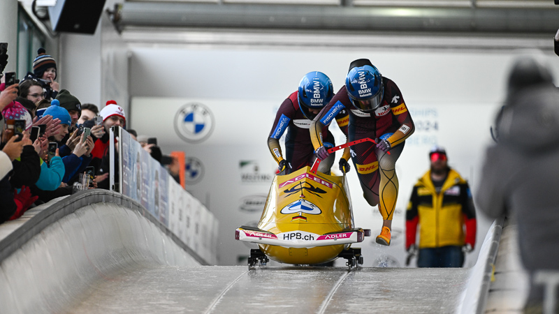 Mit Konzentration, Power und Bauerfeind zu neuen Erfolgen: Deborah Levi in Action Zwei Bobfahrer spirnten auf einer Eisbahn nach vorn. Sie sind von vorn abgebildet und von Fans an den Bobbahn-Rändern umgeben.