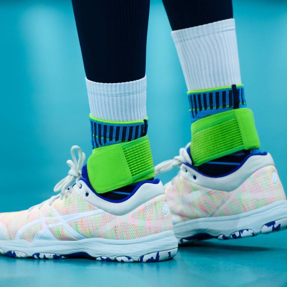 Sprungkraft mit Sicherheit – optimaler Halt für intensive Matches! Nahaufnahme der Beine eines Volleyballspielers auf dem Hallenboden. Die Bauerfeind Sports Ankle Support Rivera ist über hohen weißen Socken getragen.