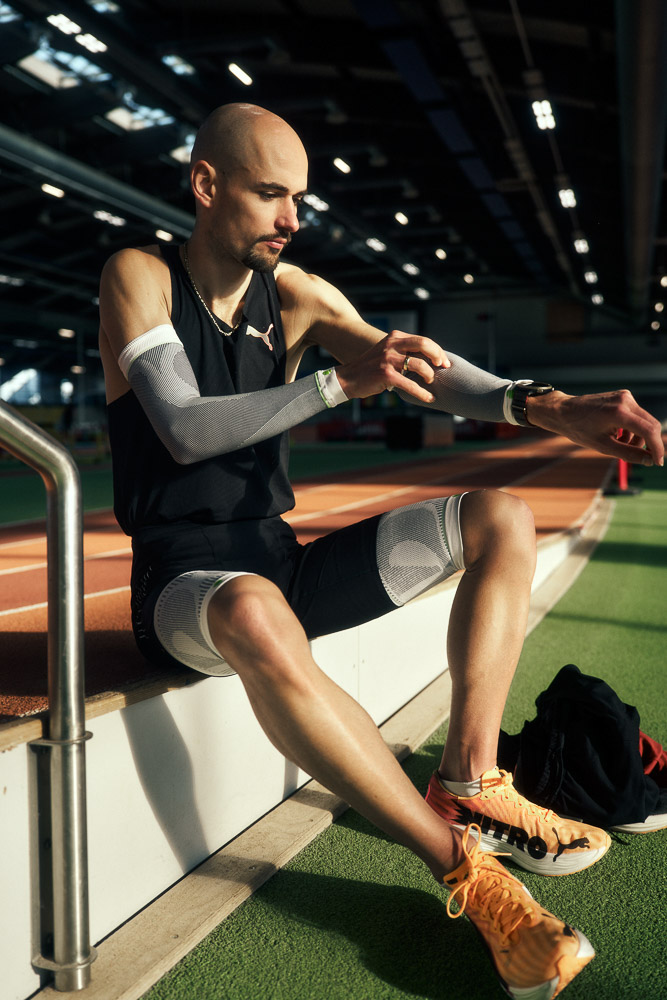 Marathonläufer Hendrik Pfeiffer sitzt am Rand einer Indoor-Laufbahn und zieht sich Kompressions-Arm-Sleeves über, als Teil seiner Wettkampfvorbereitung.