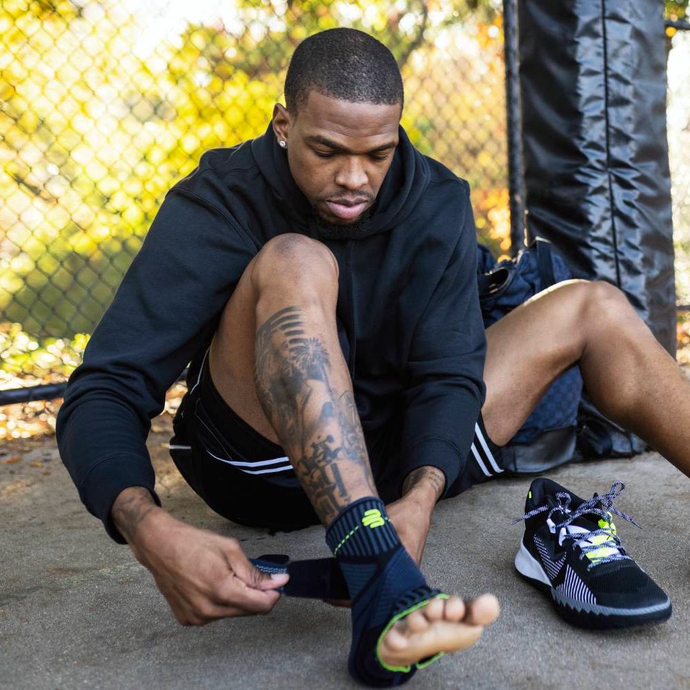 Unterstützung, wo sie zählt – Vorbereitung auf die Action! Ein Sportler sitzt im Freien und legt sich eine Bauerfeind Sports Ankle Support Bandage um den Knöchel. Seine Sportschuhe stehen bereit, und im Hintergrund ist ein Metallzaun zu sehen.