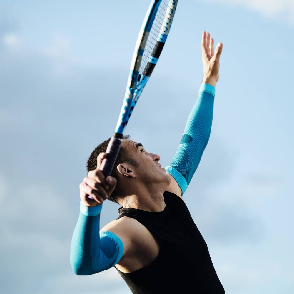Ein Tennisspieler mit blauen Kompressionsärmeln beim Aufschlag. Der Himmel ist blau mit wenigen Wolken.
