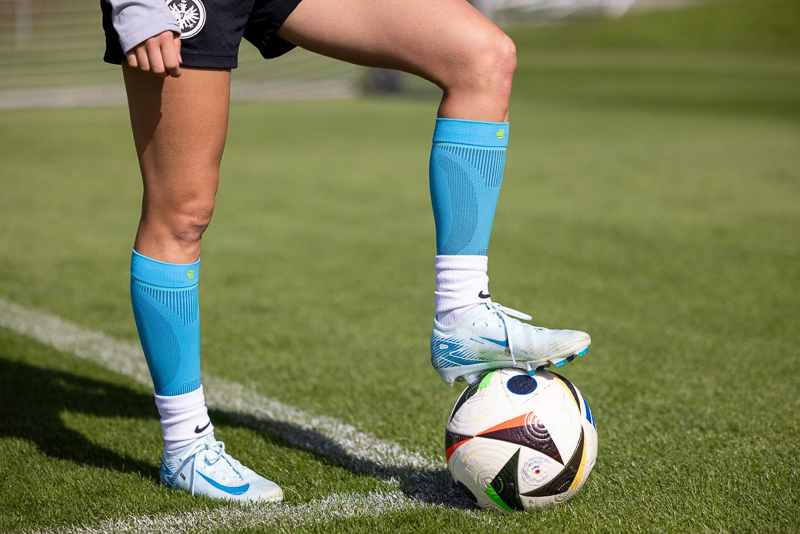 Präzision und Kontrolle mit Bauerfeind-Sports-Kompression Nahaufnahme eines Fußballspiels, bei dem eine Spielerin mit Bauerfeind-Kompressionsstrümpfen auf einem Fußball steht. Der grüne Rasen und die scharfe Darstellung des Balls und der Strümpfe betonen die sportliche Leistung und die Qualität der Ausrüstung.