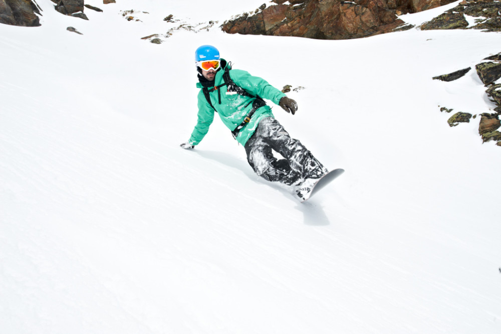 Snowboarder in grüner Jacke ist an einer extrem steilen Stelle hinter einem Felsen mit der hinteren Hand im Schnee und schaut in Richtung Tal