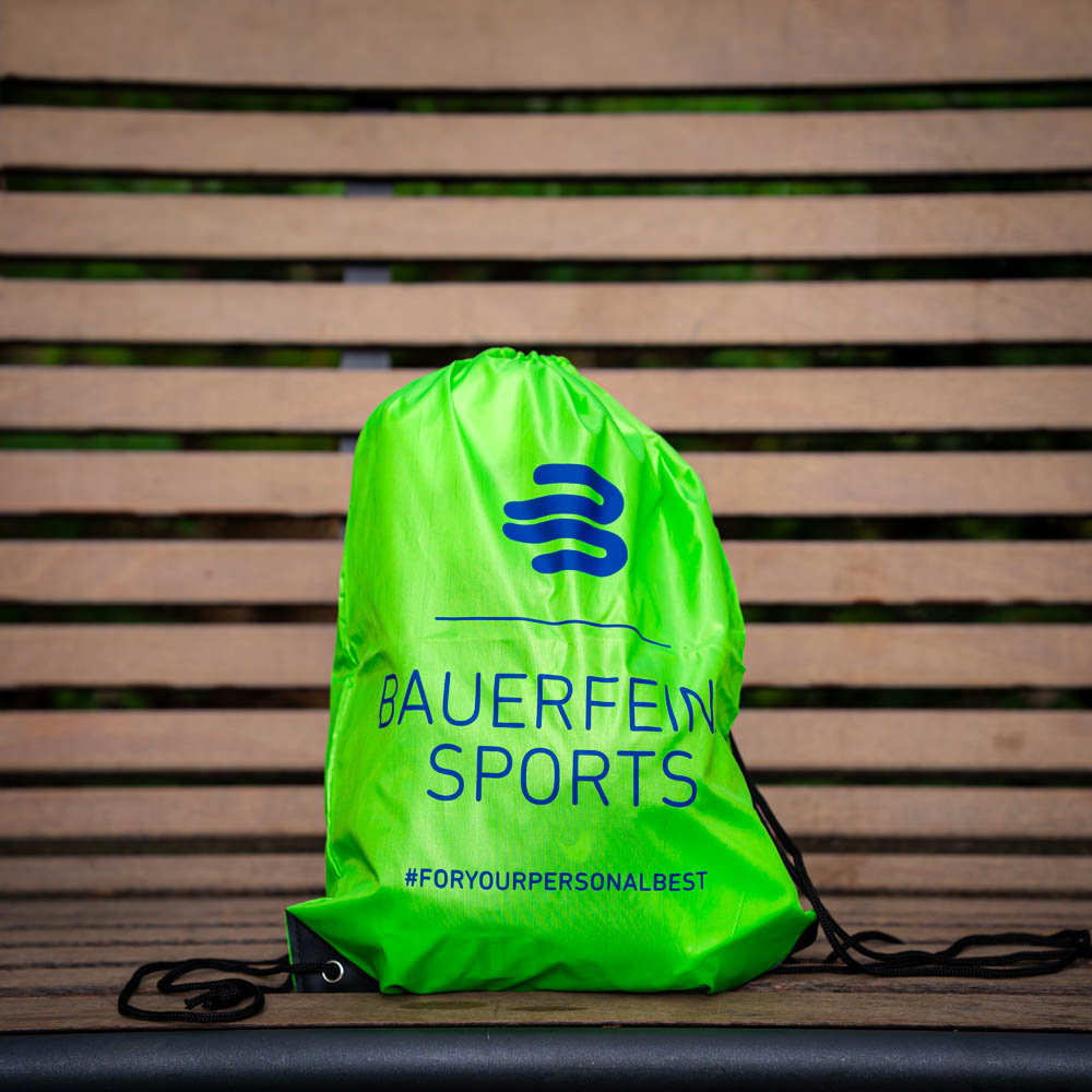 Praktisch und stylisch: Der Bauerfeind Sports Turnbeutel für deine Ziele Ein grüner Bauerfeind Sports Turnbeutel liegt auf einer Holzbank, gut sichtbar mit dem Logo und dem Hashtag #foryourpersonalbest.