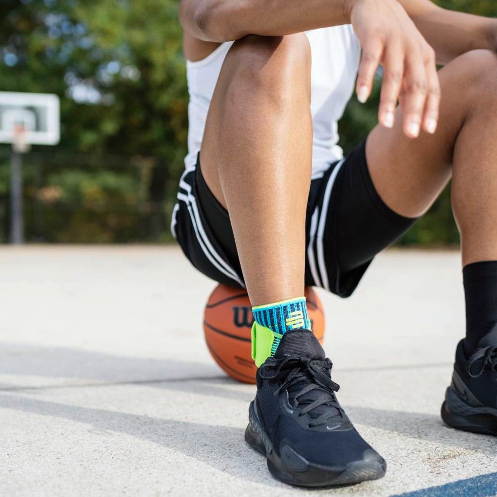 Stabil auf dem Court – mit sicherem Halt! Ein Basketballspieler sitzt auf dem Spielfeld mit einem Basketball unter dem Bein. Am Knöchel trägt er die Bauerfeind Sports Ankle Support Rivera in Blau-Grün.