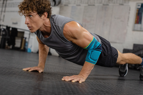 Junger Mann mit Ellenbogenbandage macht Push-ups auf dem Boden eines Trainingsraums