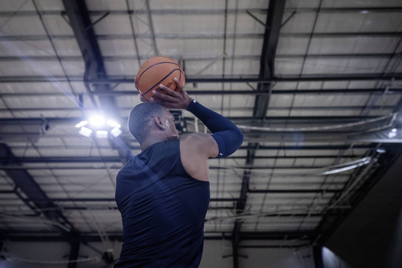 Basketballspieler wirft Ball und trägt Armsleeve Basketballer mit blauem Armsleeve in Sporthalle setzt mit den Händen oberhalb des Kopfes zum Wurf an