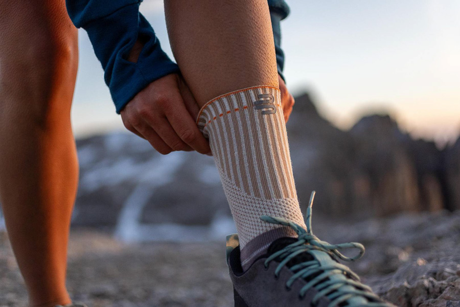 Ein Wanderer passt seine spezielle Outdoor-Bandage mit verstärktem Bündchen an. Im Hintergrund sind Berge und ein Sonnenuntergang zu sehen.