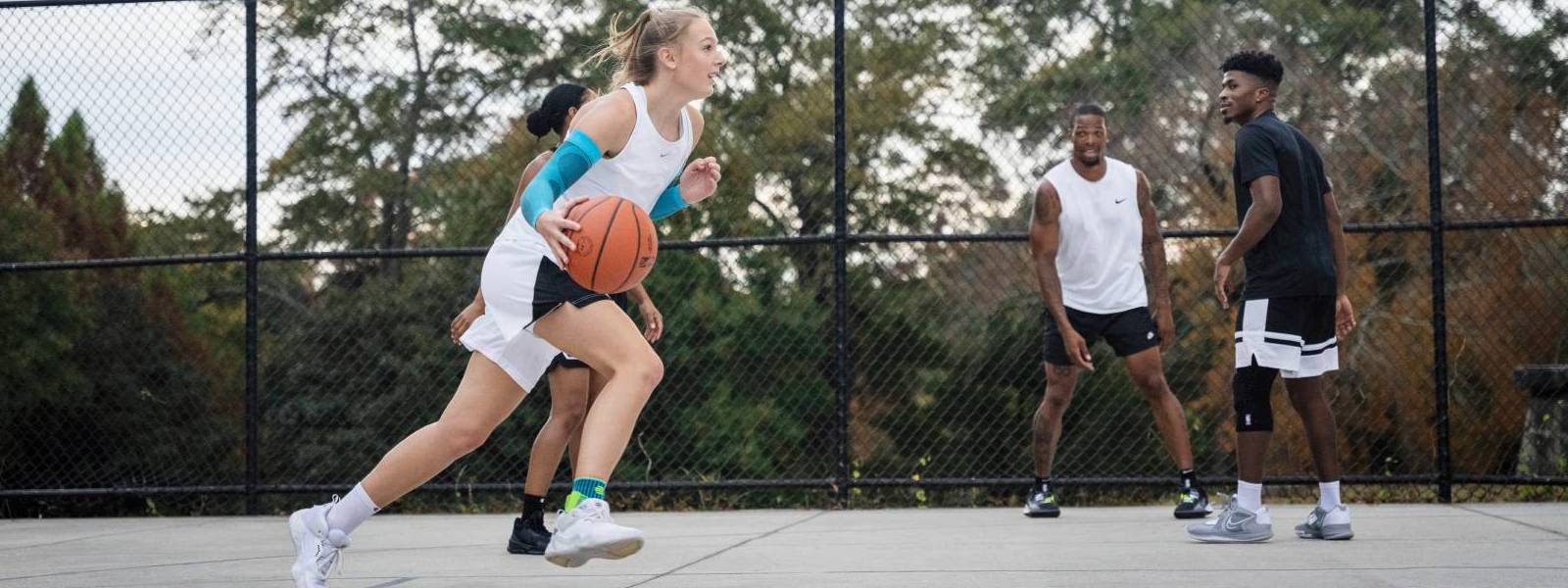 Basketball Spielerin dribbelt auf einem Freiplatz an ihren Gegenspielern vorbei und trägt dabei blaue Arm Sleeves