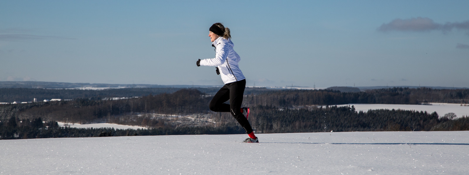 Frau in weißer Laufjacke joggt durch gleichmäßiges Schneefeld	 im Hintergrund wechseln sich kleine Wälder und Lichtungen ab