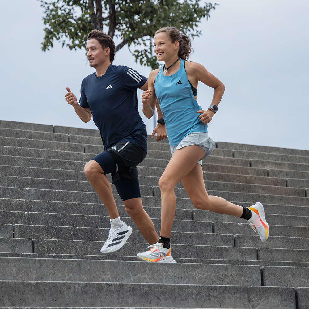 Eine sportliche Frau und ein Mann laufen gemeinsam dynamisch eine breite Steintreppe hinunter. Beide tragen Sportkleidung und Schuhe von adidas.