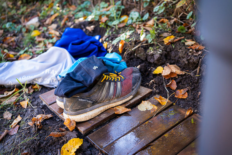 Vor dem Sprung ins Eiswasser: Schuhe sicher geparkt, Mut gesammelt! Ein Paar Laufschuhe mit gelben Schnürsenkeln steht auf einem Holzbrett. Daneben liegt ein Haufen Kleidung bestehend aus einem blauen Handtuch, einer grauen Jacke und schwarzen Handschuhen. Herbstlaub ist auf dem Boden verstreut.