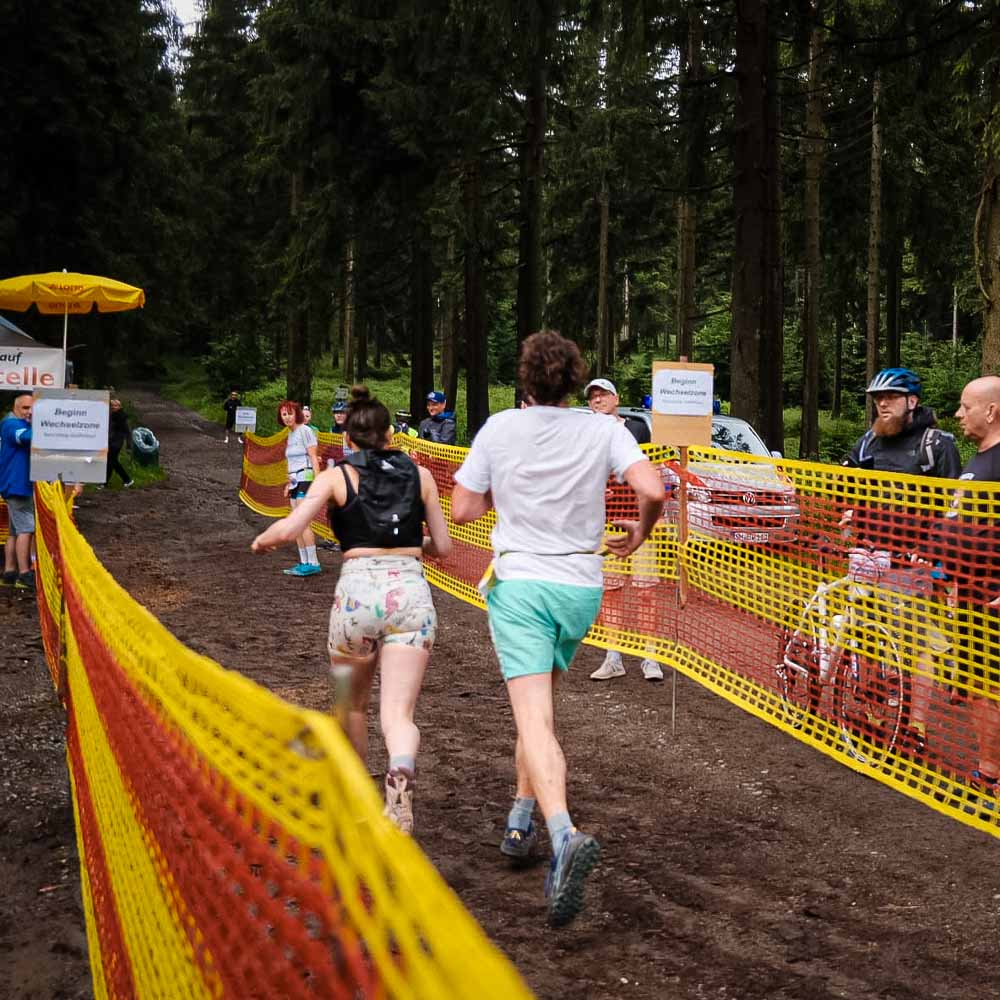 Gemischter Staffellauf durch den Rennsteig Ein Mann und eine Frau laufen durch den Wald, beobachtet von Passanten, während der Boden schlammig ist