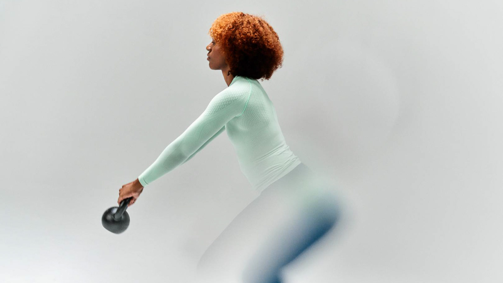 Eine Frau in einem mintfarbenen Kompressions-Sports-Wear-Shirt führt eine Kettlebell-Übung durch. Der Fokus liegt auf ihrer Bewegung und der funktionalen Sportswear vor einem neutralen Hintergrund.