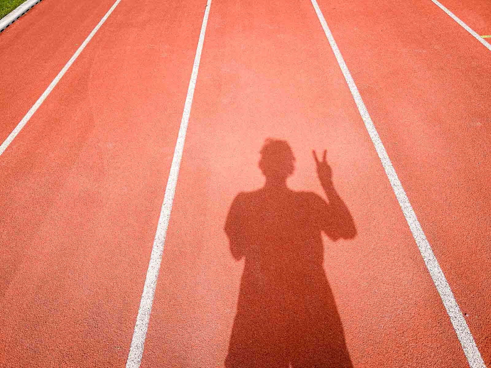 Schatten auf der Laufbahn – Erfolg und Zielerreichung nach dem Lauftraining. Der Schatten eines Läufers auf einer roten Laufbahn, der das Victory-Zeichen mit der Hand zeigt. Die Szene symbolisiert Erfolg und Zielerreichung im Lauftraining.