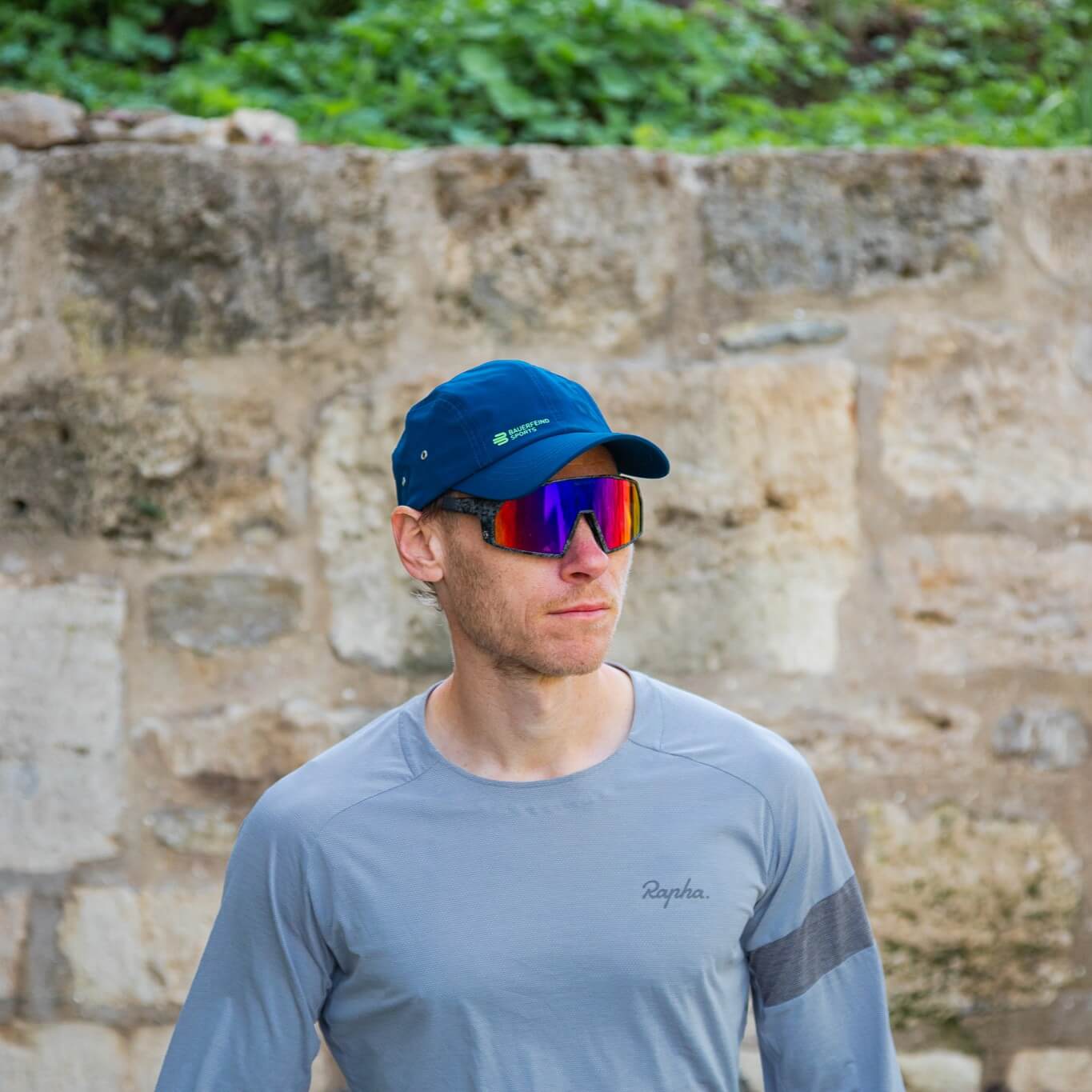 Sportler trägt eine blaue Bauerfeind Sports Cap und verspiegelte Sportbrille vor einer Steinwand – bereit für aktive Outdoor-Momente.