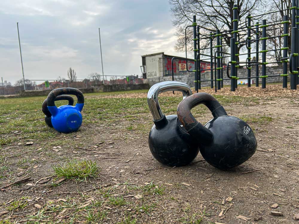 Mehrere Kettlebells stehen auf unbefestigtem Boden in einem Outdoor-Trainingspark, im Hintergrund ist ein Calisthenics-Gerüst zu sehen. Das Bild zeigt ein funktionelles Setup für HYROX-Training ohne Gym