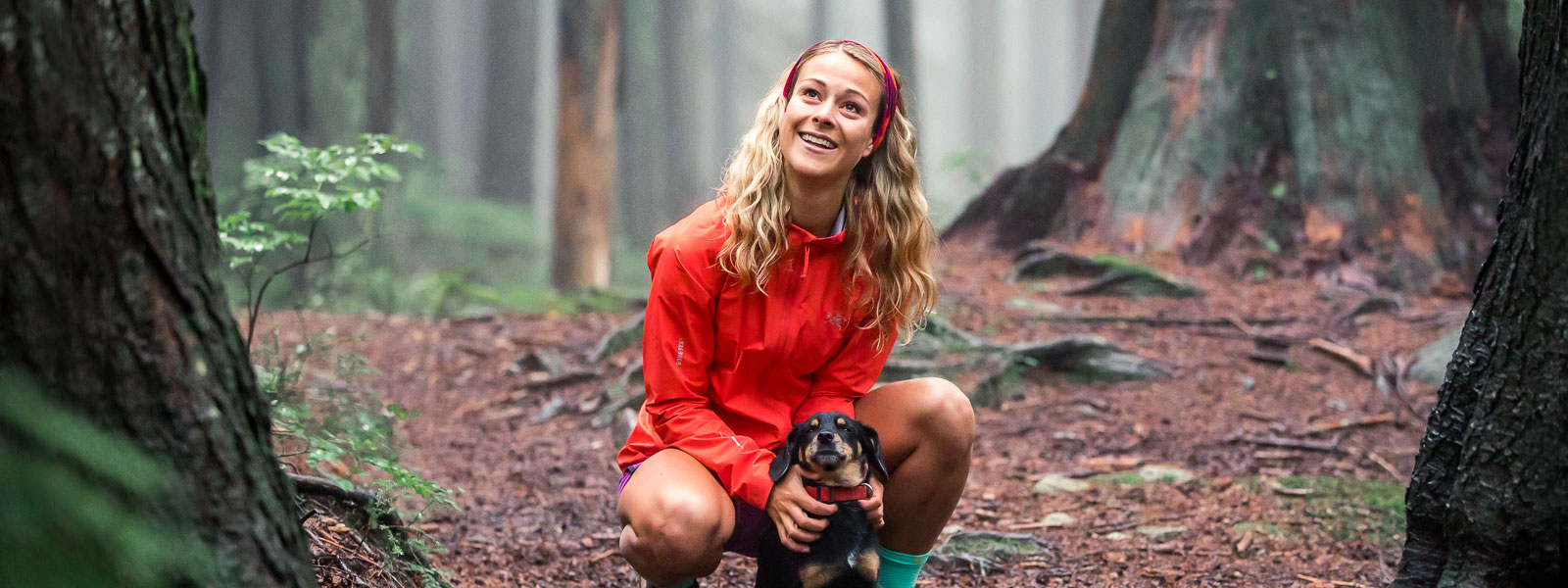 Frau mit roter Laufjacke und Stirnband hält ihren kleinen Hund auf einem Waldweg und lächelt