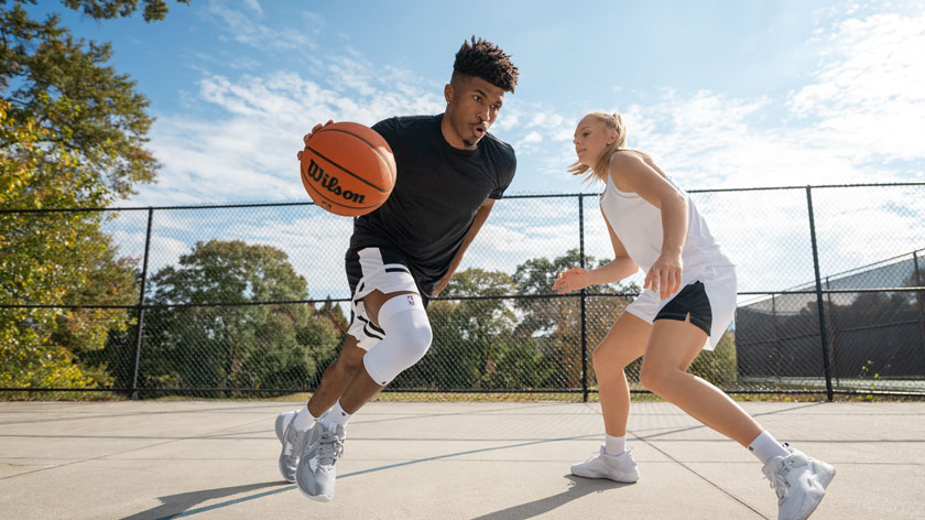 Ein Basketballspieler treibt den Ball energisch über einen Freiplatz, verfolgt von einer Gegnerin. Er trägt eine weiße Kniebandage, die seinen Bewegungsapparat unterstützt.