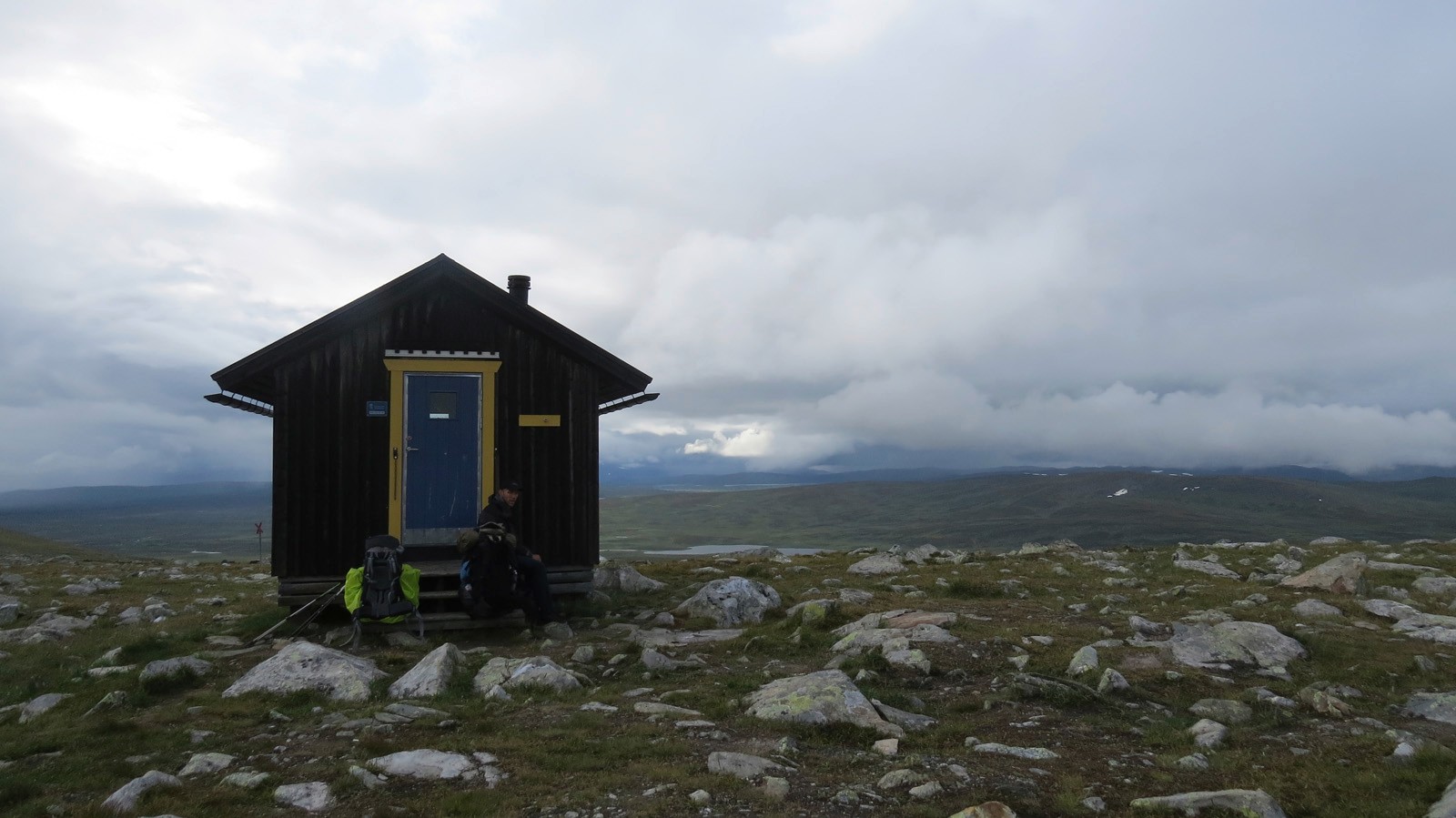 Eine einsame Hütte im Gebirge inmitten von begrasten Wiesen mit kleinen Felsen	 im Hintergrund die dichte Wolkendecke und ein kleiner Bergsee