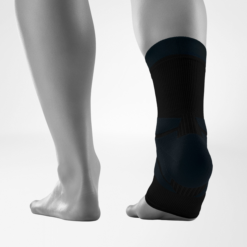 Rückansicht beider Füße vor neutralem Hintergrund, wobei ein Fuß mit dem schwarzen Outdoor Compression Ankle Support ausgestattet ist. Die Knöchelbandage umschließt den Sprunggelenkbereich eng anliegend.