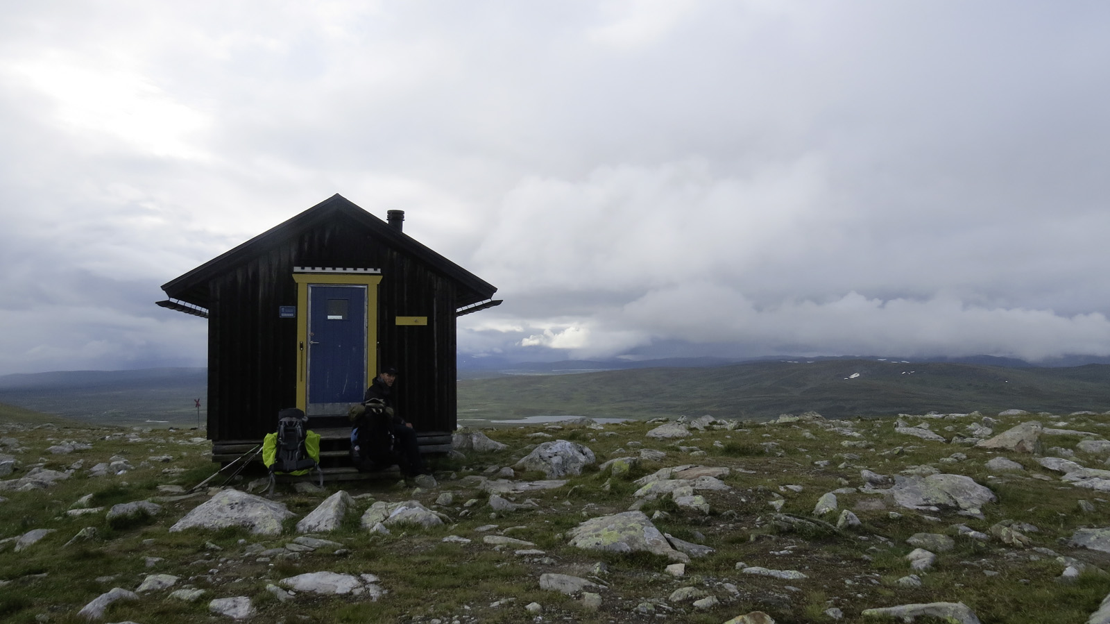 Eine einsame Hütte im Gebirge inmitten von begrasten Wiesen mit kleinen Felsen	 im Hintergrund die dichte Wolkendecke und ein kleiner Bergsee